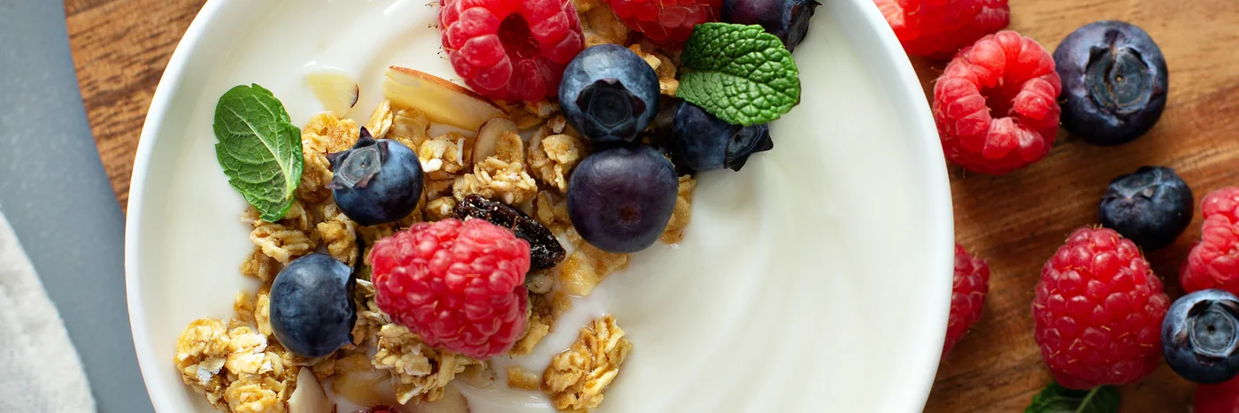 A bowl of plain yoghurt topped with granola, raspberries and blueberries on a dark table.