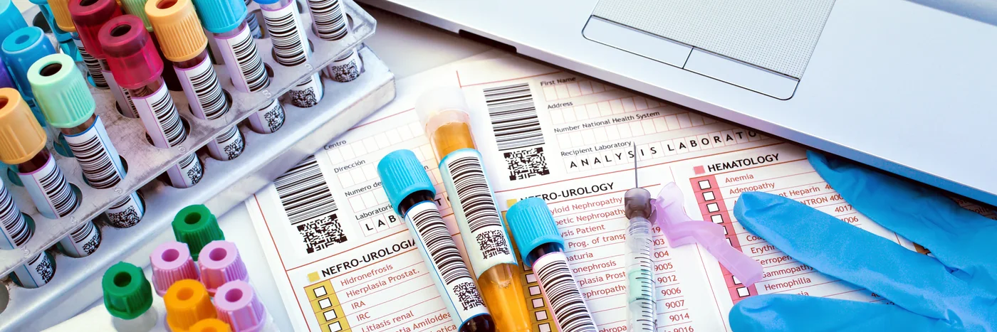 A laboratory bench with racks of sample tubes, barcoded requisition forms and a technician’s gloved hand.