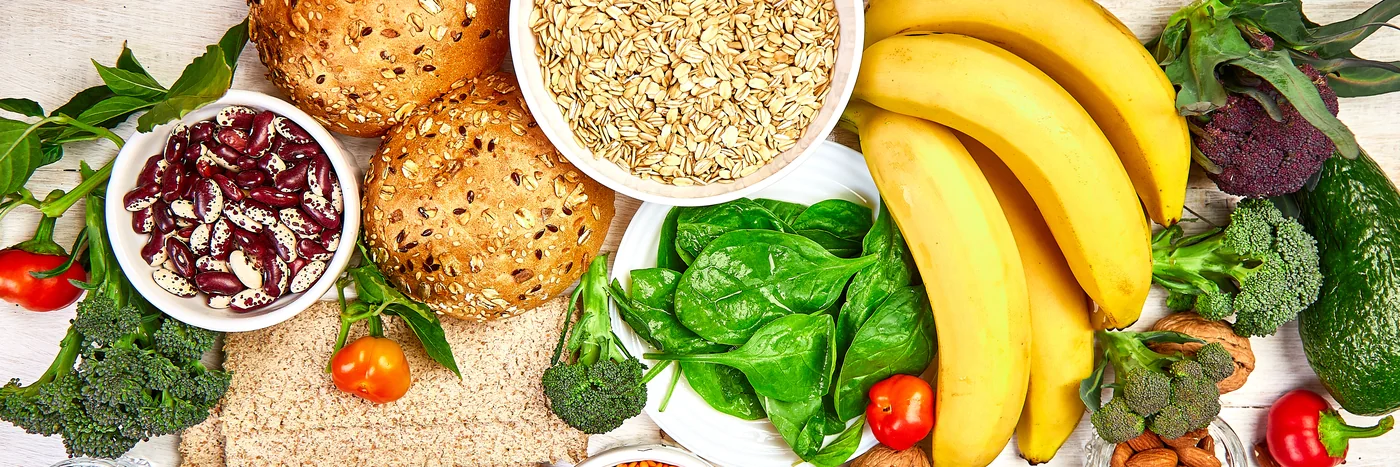 An overhead spread of high-fibre foods on pale wood: oats, seeded rolls, beans, spinach, broccoli, bananas and berries.